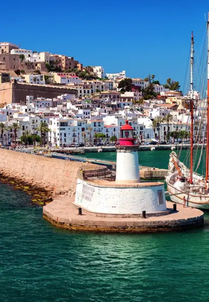 Küstenstadt mit Festung, Hafenpromenade und Segelschiff vor türkisblauem Meer.