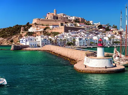 Küstenstadt mit Festung, Hafenpromenade und Segelschiff vor türkisblauem Meer.